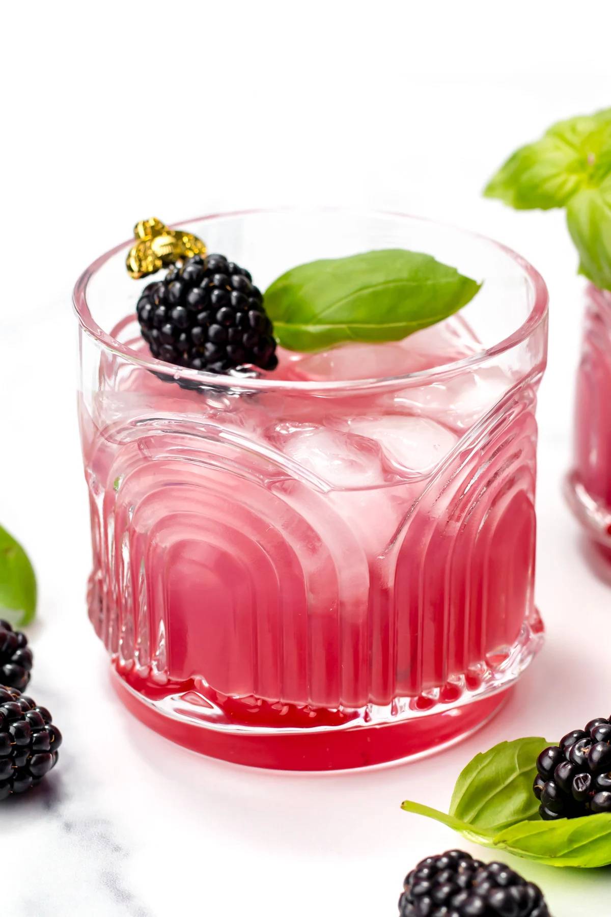 A deep pink drink is garnished with a blackberry and herb leaf on a gold skewer.