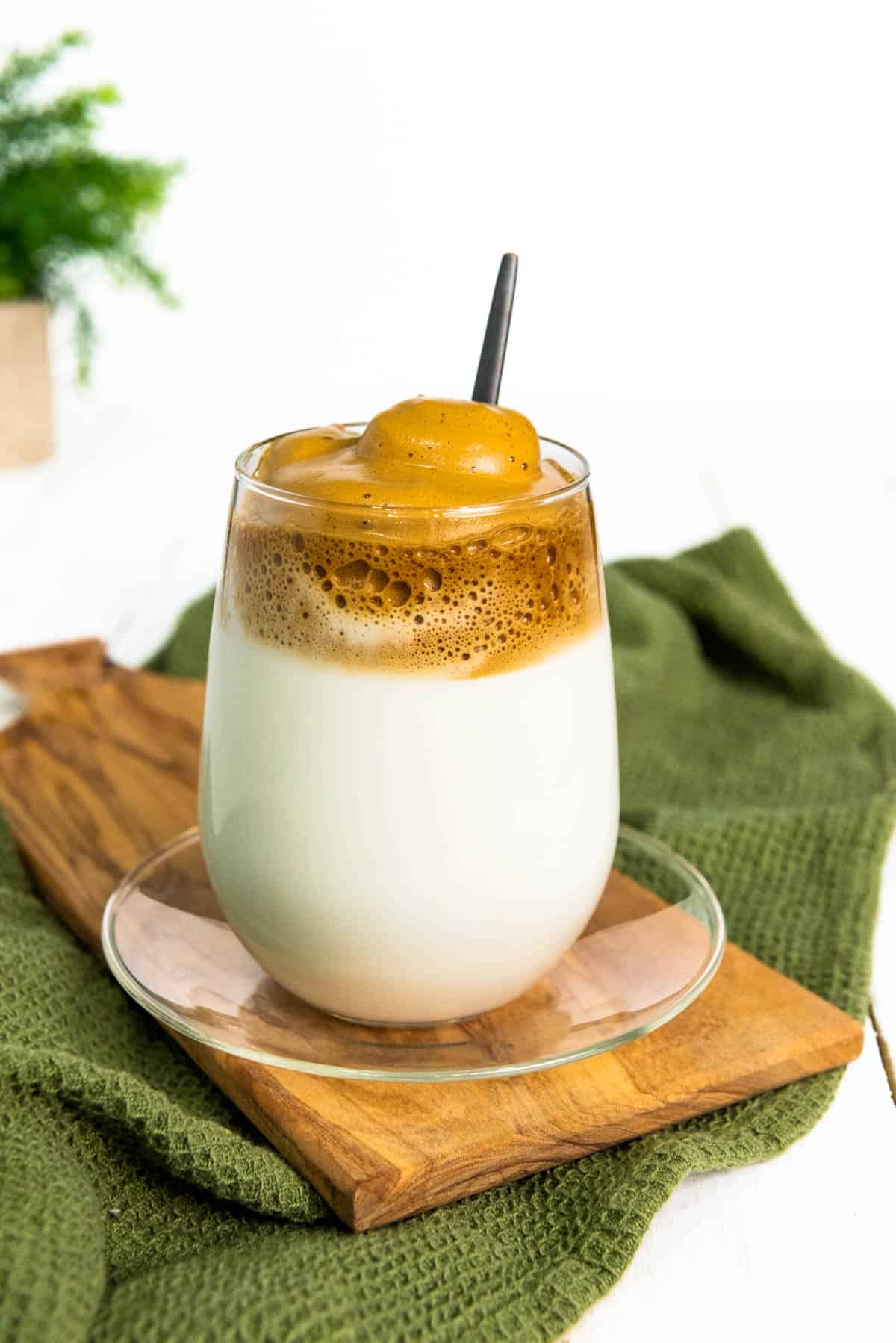 A whipped coffee drink in a stemless glass on a clear glass plate and a wooden cutting board.