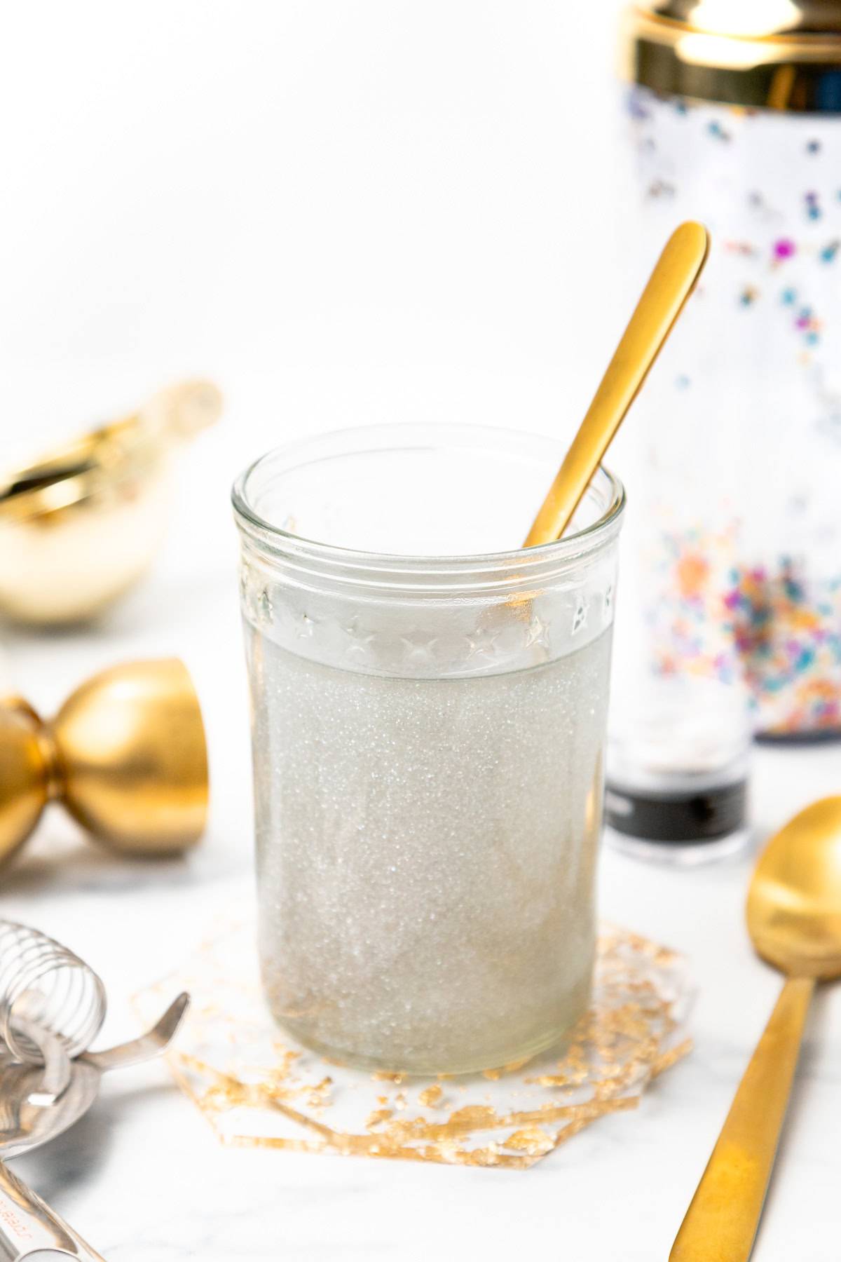A glass jar of sparkling syrup sits with a gold spoon in it.