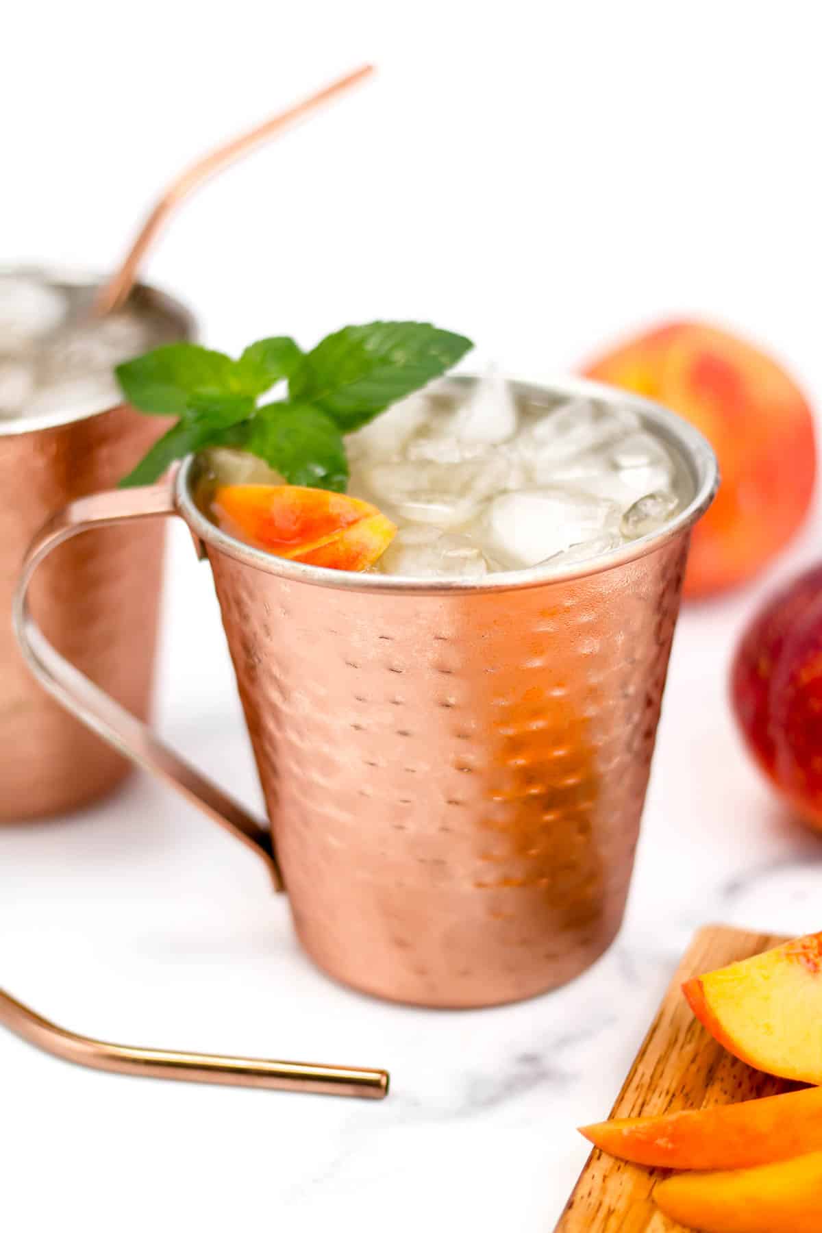 A copper mug holds an icy beverage garnished with peach slices and an herb sprig.