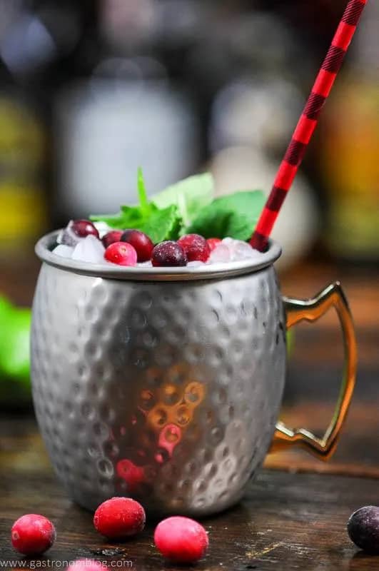A silver mule mug topped with cranberries, a mint sprig and a red and maroon striped straw.