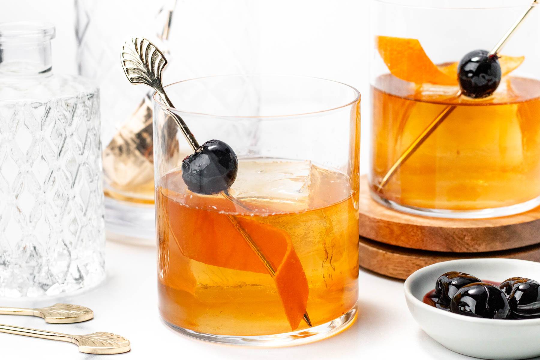 An amber cocktail in a rocks glass with a gold cocktail pick holding a cherry.
