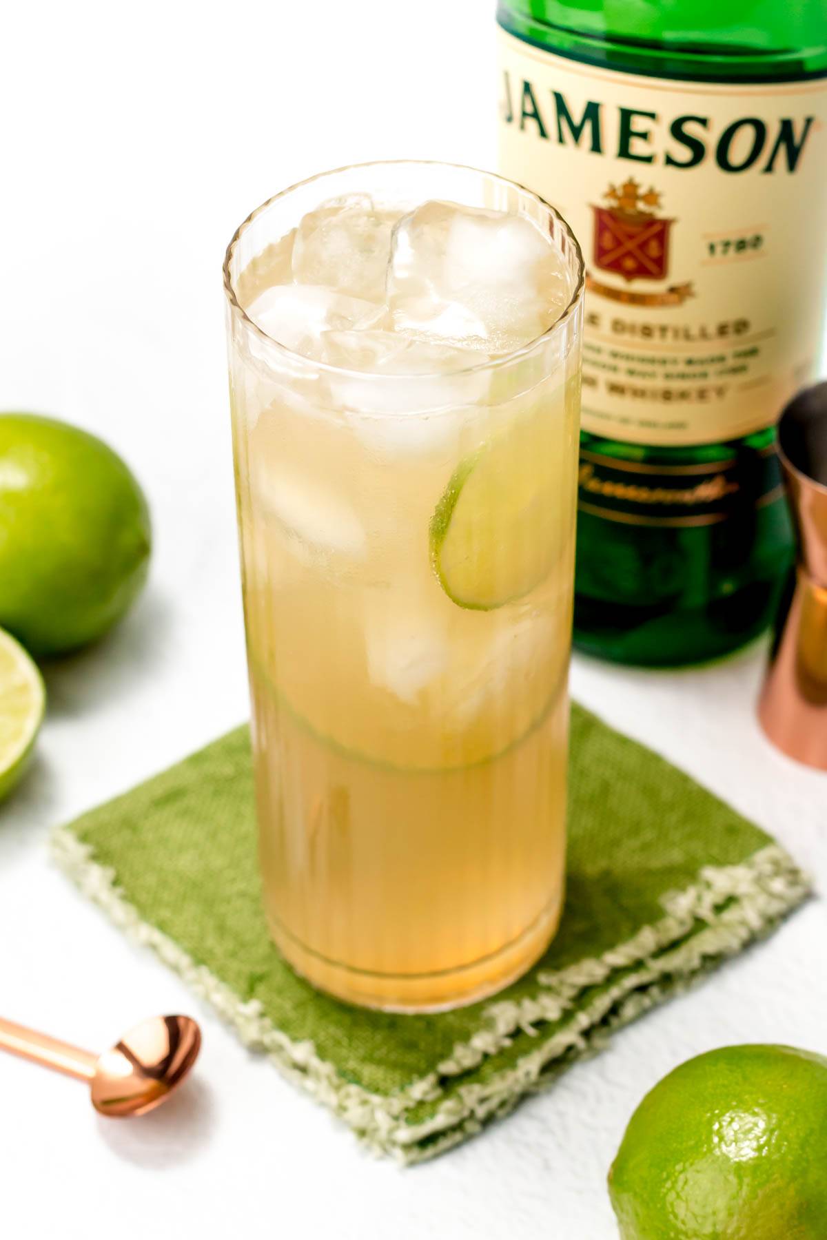 A tall glass holds a pale yellow drink on a green fabric coaster near a bottle of Jameson.