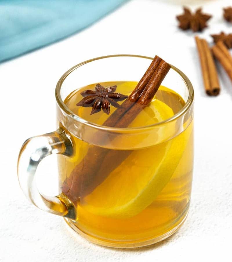 A clear glass mug holds an amber liquid with a cinnamon stick, star anise and lemon round.