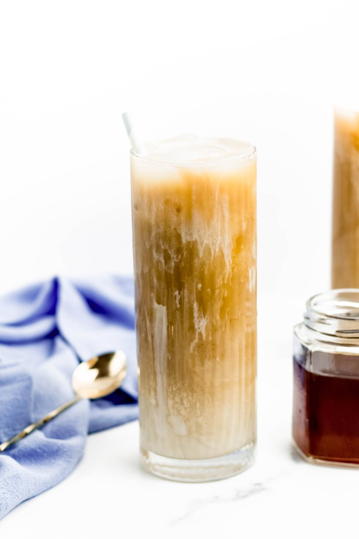 A tall glass holds a coffee drink near a jar of brown syrup and a gold spoon.