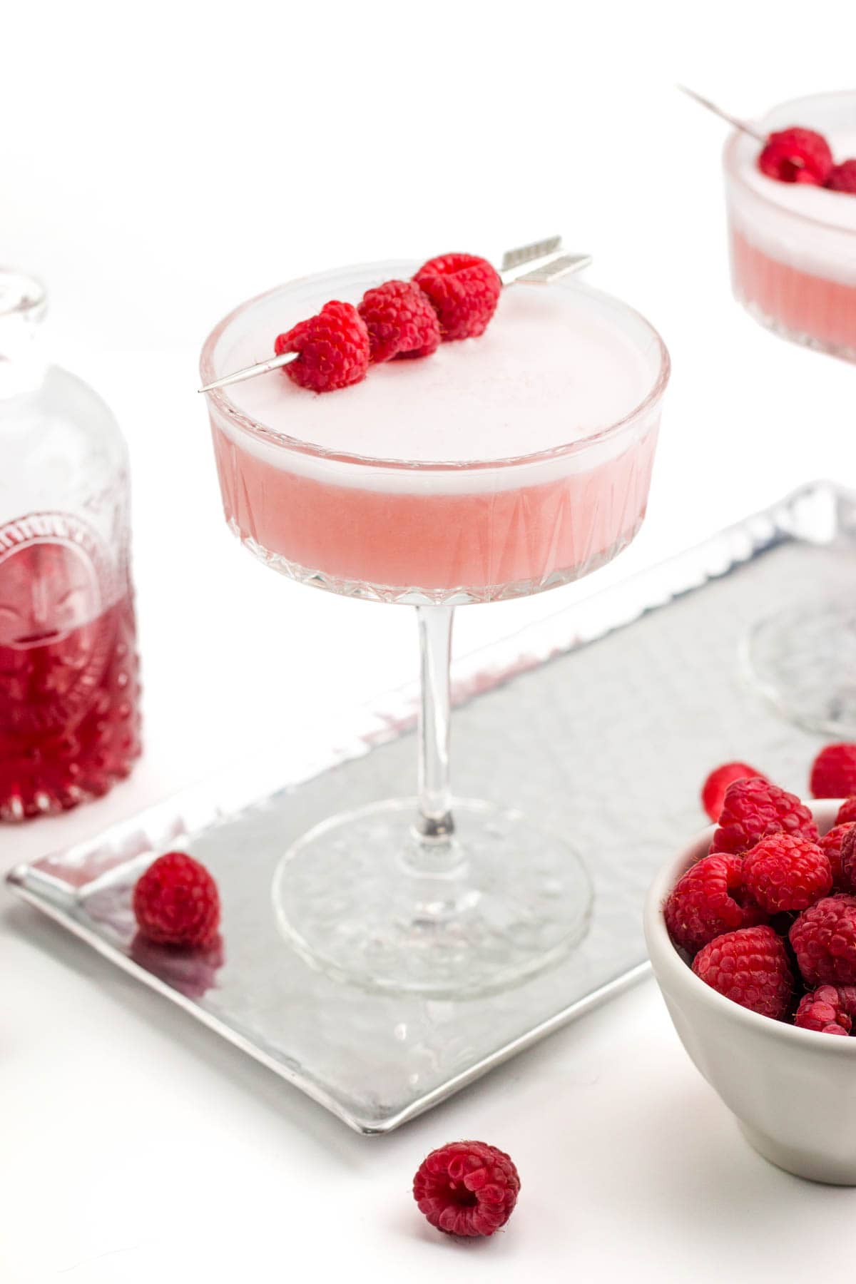 A pink cocktail garnished with raspberries on a skewer.