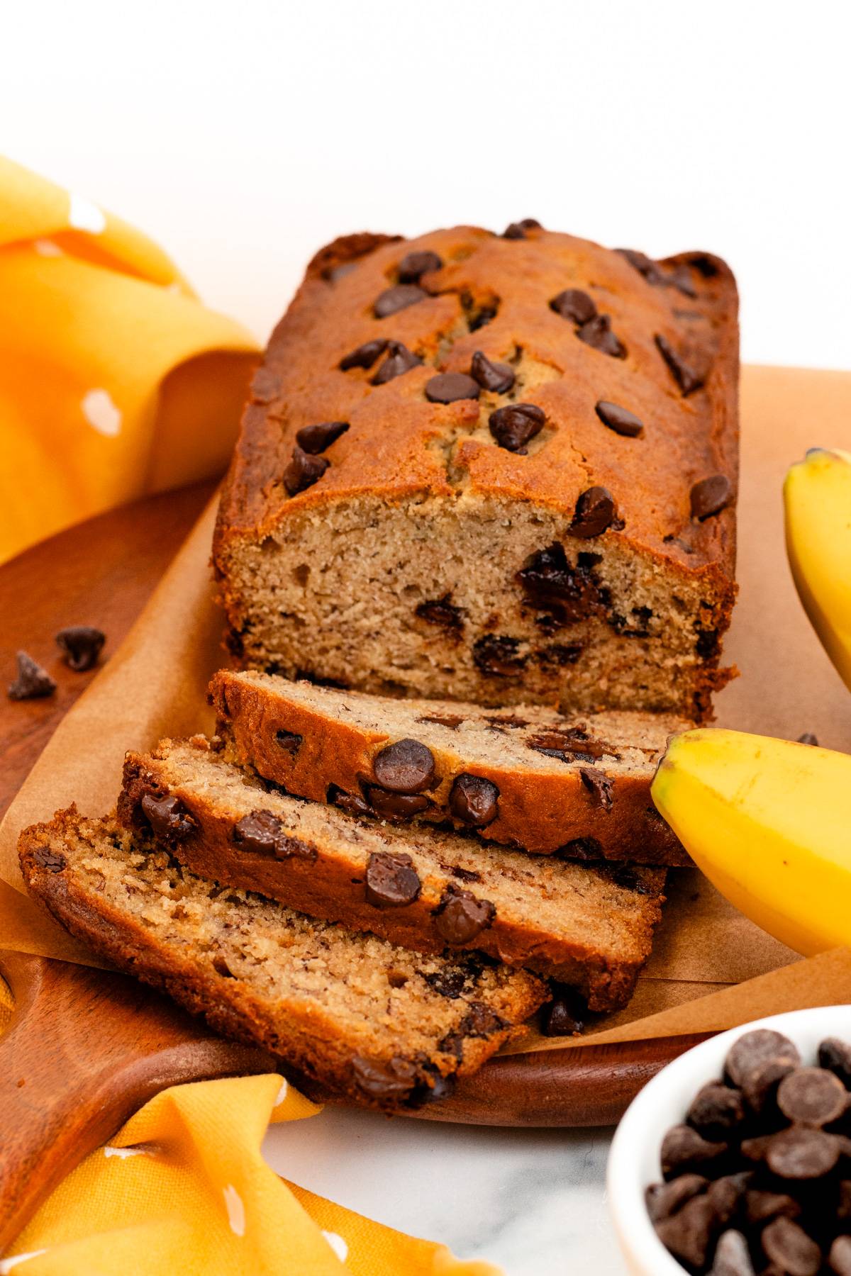 A bread filled with chocolate chips sliced on a brown wooden board near bananas.