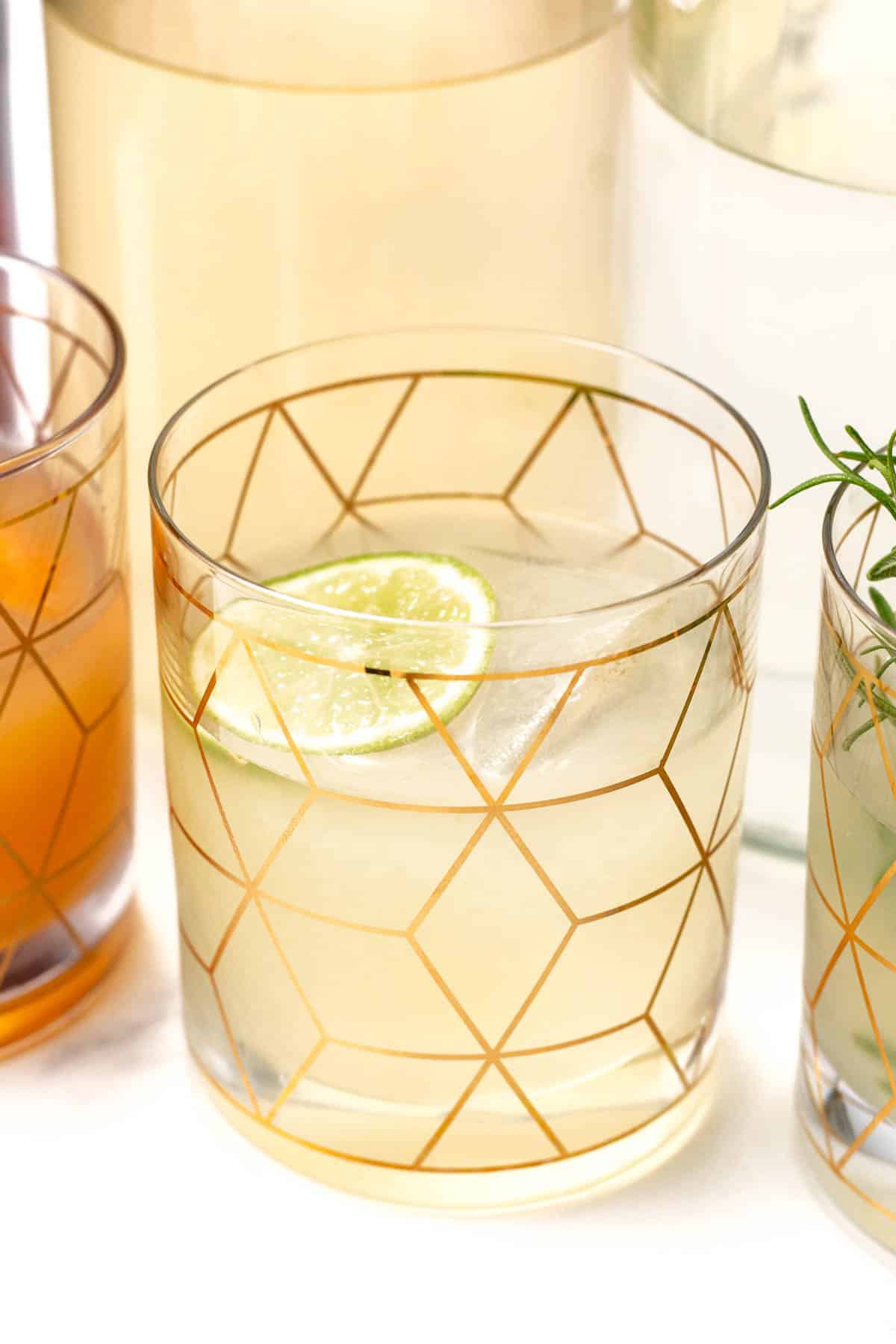 A glass with a gold geometric pattern holds a pale yellow drink with a lime wheel.