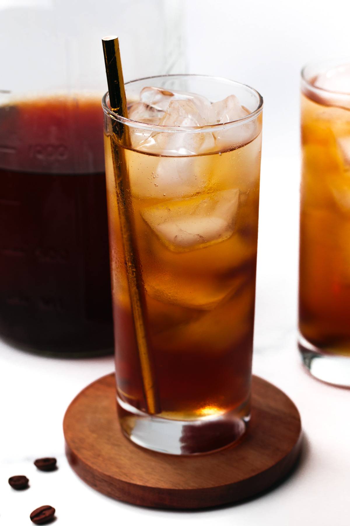A brown beverage sits on a round wooden coaster with a bronze straw sits near a carafe of coffee.