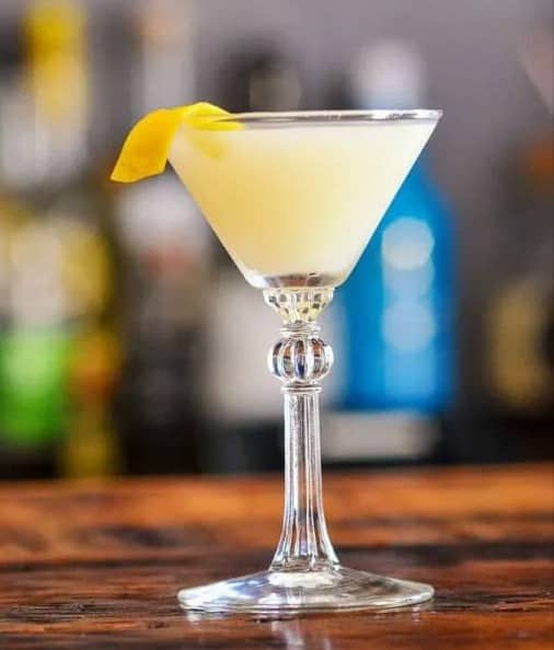 A stemmed glass holding a yellow drink with a lemon twist on the rim.