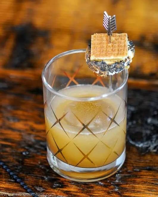 A rocks glass holds a drink garnished with a small s'mores on a skewer.