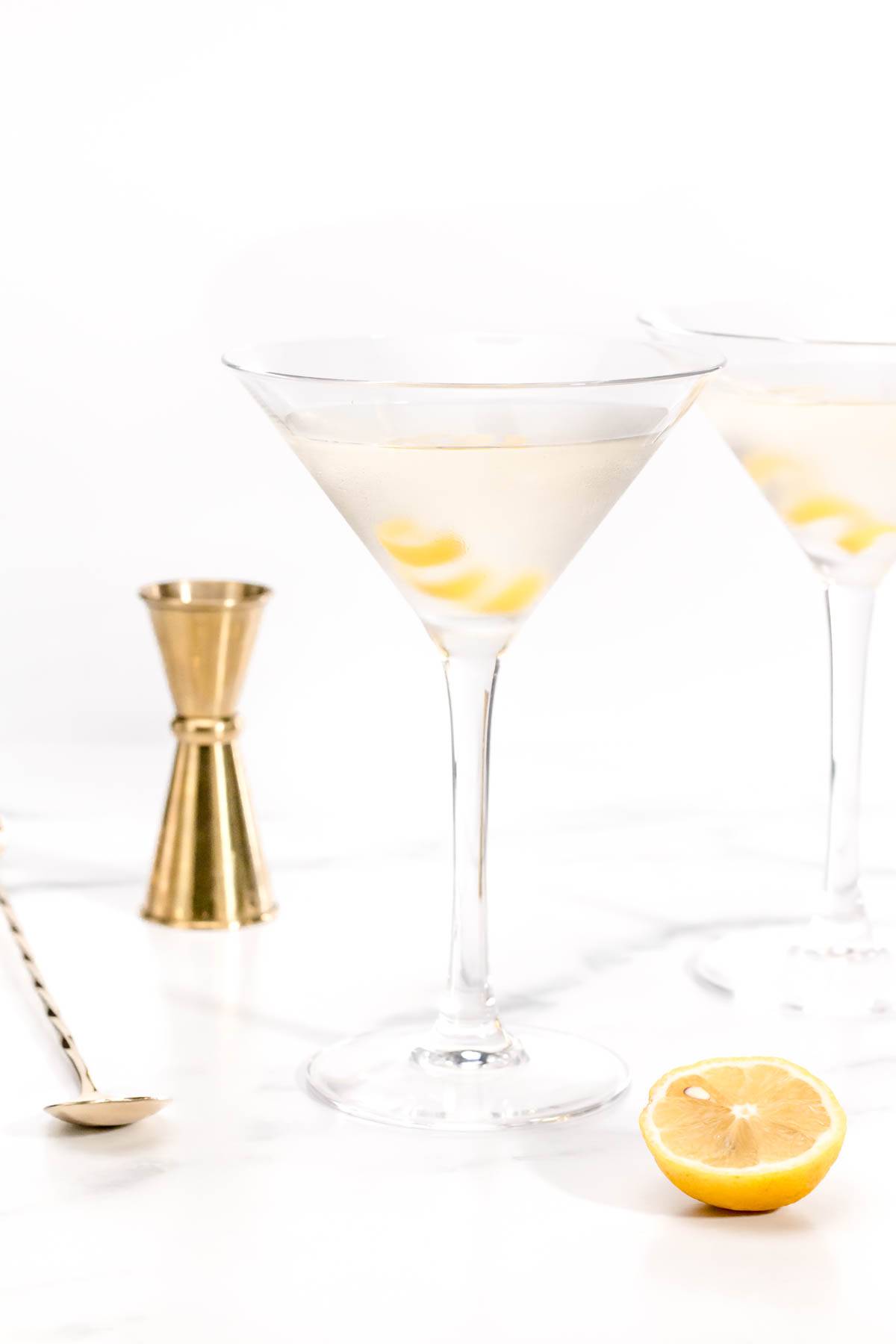 Two clear martinis with lemon twists sit on a marble surface alongside gold bar tools and half a lemon.