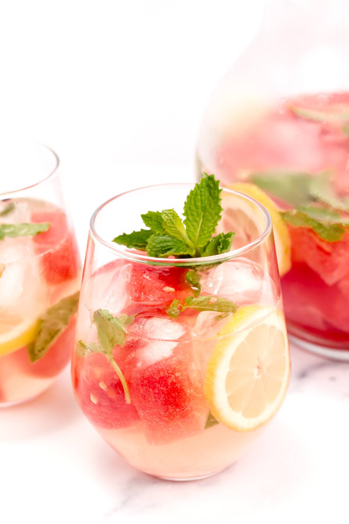 A glass of white wine sangria with watermelon chunks, lemon rounds and mint sprigs.