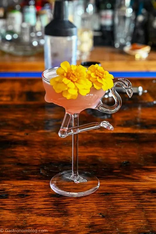 A flamingo glass holds a pink drink with orange flowers as a garnish.