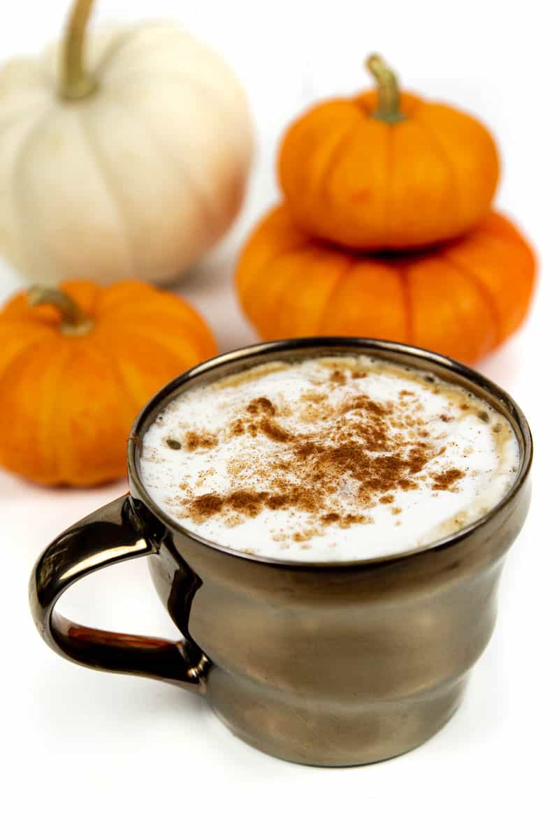 A latte in a shiny bronze cup topped with cinnamon near small white and orange pumpkins.
