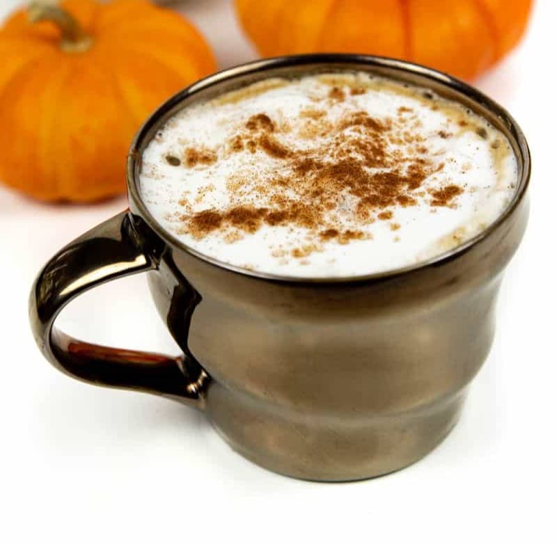 A bronze mug holds a coffee drink topped with foam and cinnamon near small pumpkins.