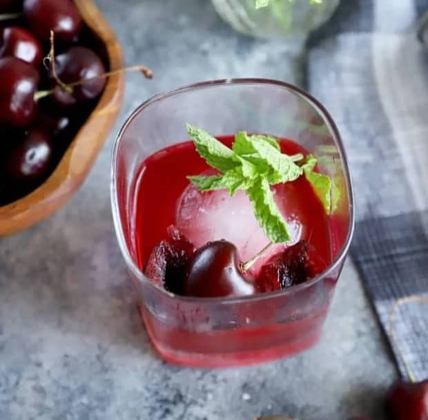 Overhead view of a deep red drink with cherries and a sprig of mint.