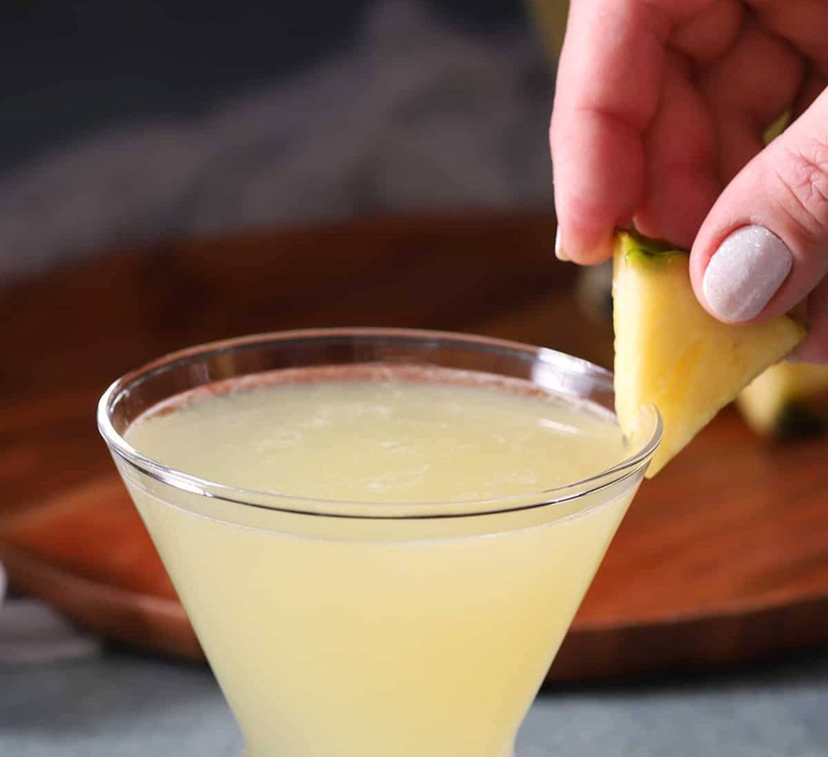 A hand places a slice of pineapple on the rim of a martini glass that holds a pale yellow drink.