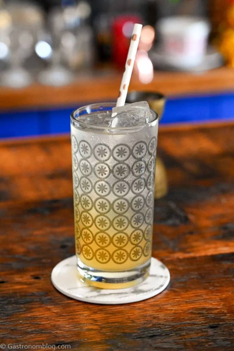 A tall glass with silver detailing holds a pale beverage with a white paper straw.