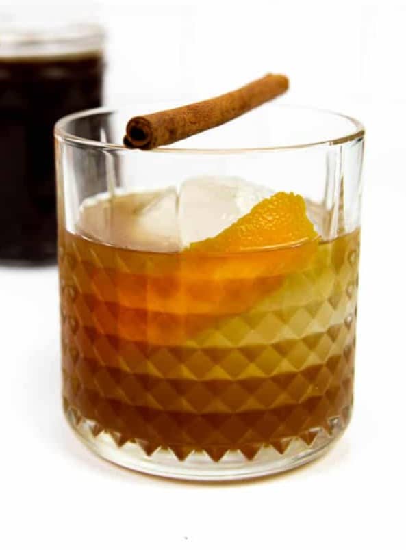 A rocks glass with amber liquid and an orange peel topped with a cinnamon stick.