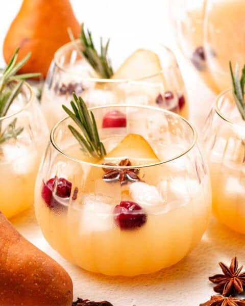 Pale orange punch sits in multiple cups garnished with berries and herbs.