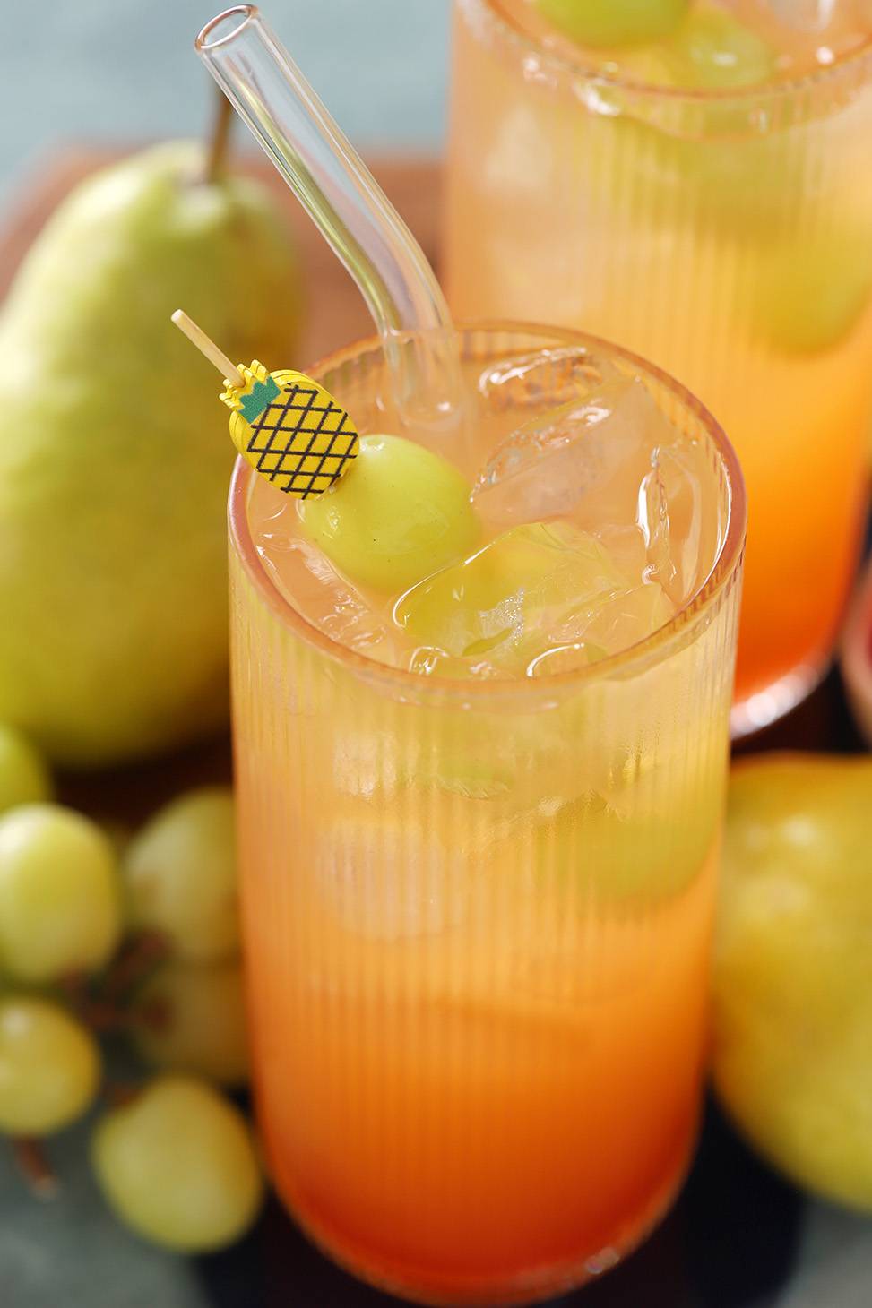 An orange drink with a grapes on a wooden pineapple skewer sits in front of pears and green grapes.