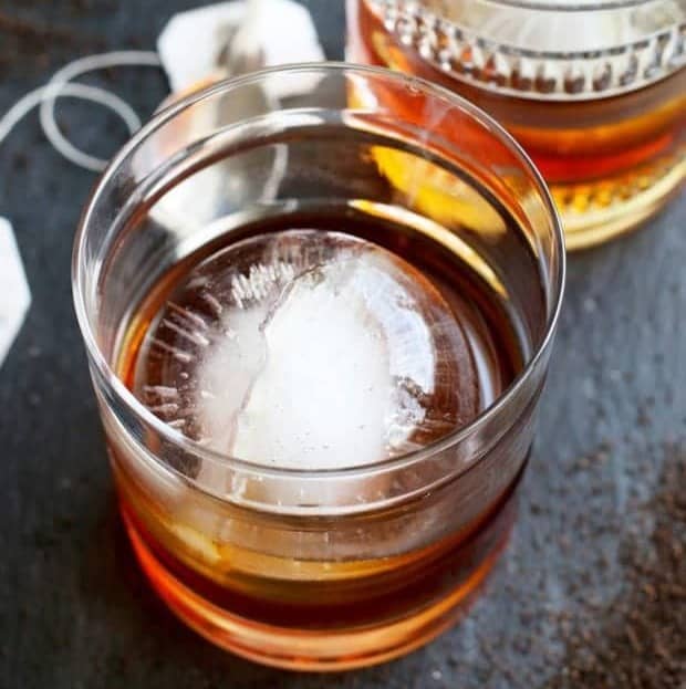A brown whiskey drink with a sphere ice cube in a rocks glass next to a tea bag. 