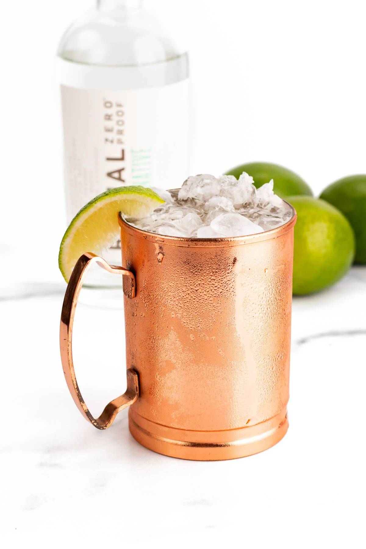 A copper mug holds a cocktail with crushed ice and a lime slice on the rim near a bottle of non-alcoholic vodka.