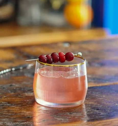 A short glass with a gold rim holds a light pink cocktail garnished with a cocktail skewer of cranberries resting across the drink.