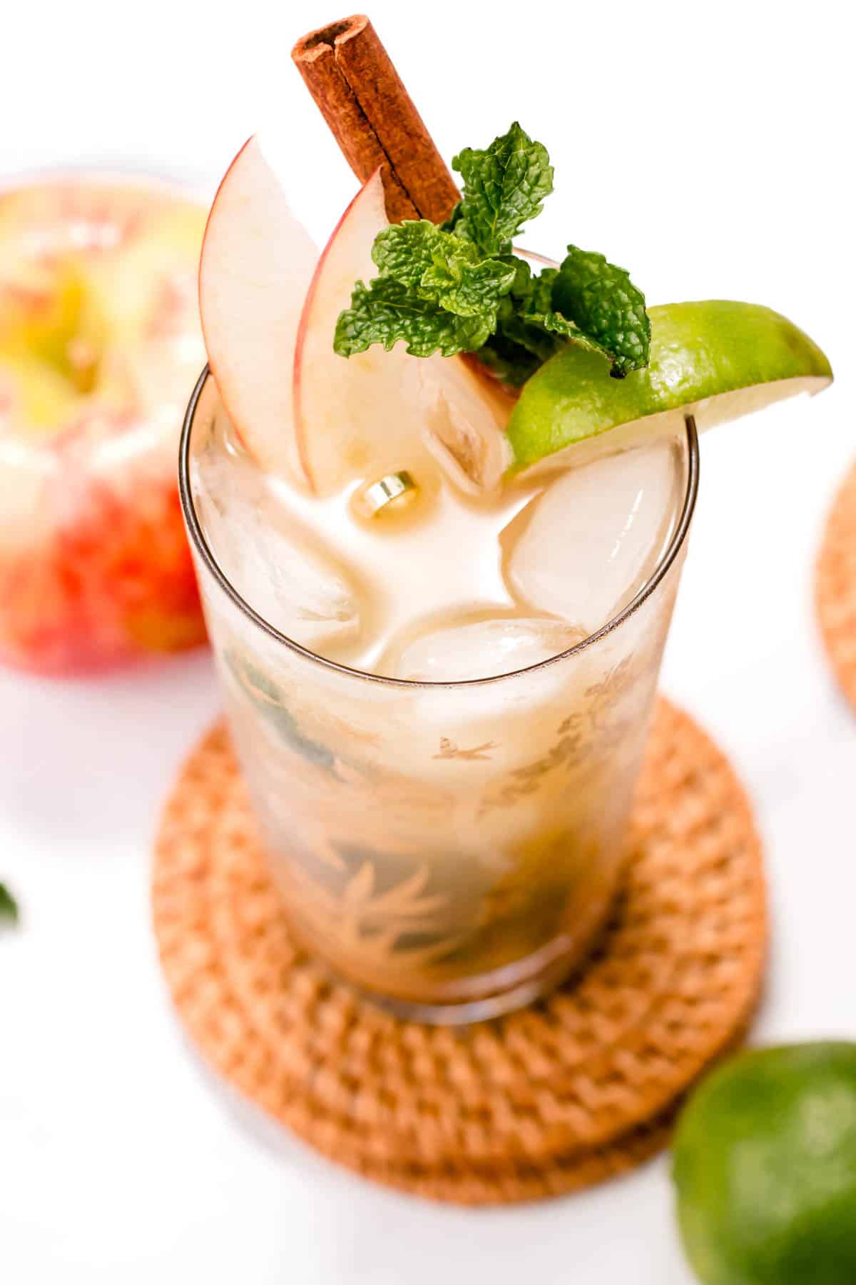 A cocktail sits on a round rattan coaster garnished with apple slices and an herb sprig.