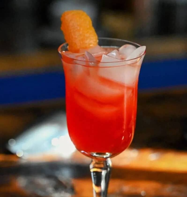A red drink in a stemmed glass with an orange fruit garnish.