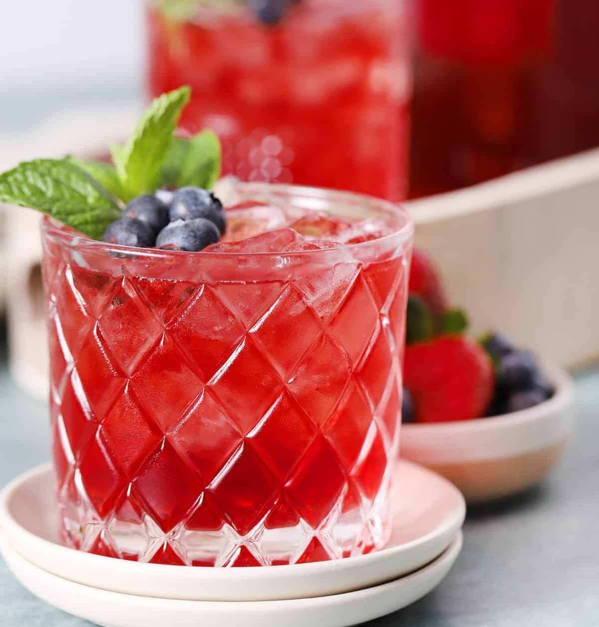 A red drink in a glass topped with blueberries and a mint sprig.