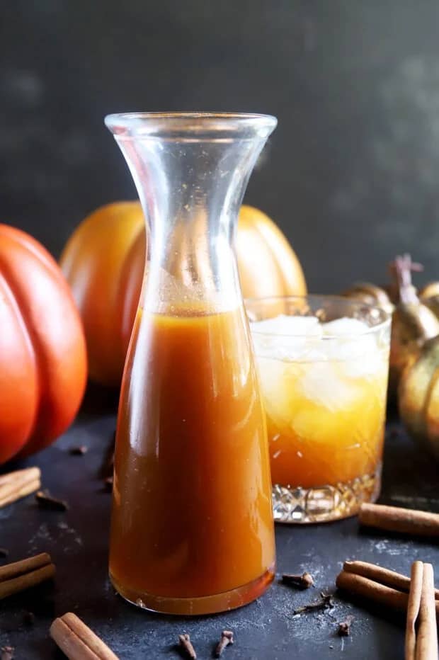 A glass carafe holds amber simple syrup near a glass with a light orange drink and some fake orange pumpkins.