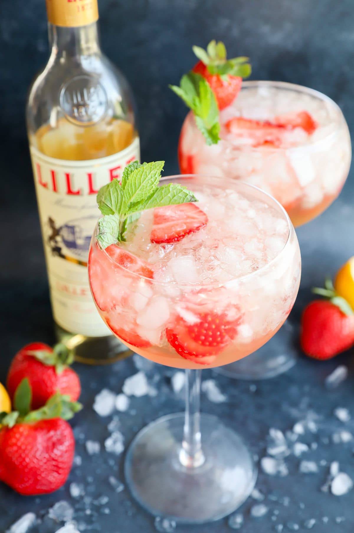 A stemmed glass holds a pale pink drink with strawberry slices and a mint sprig near a bottle of Lillet Blanc.