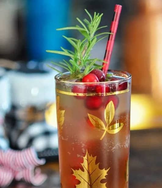 A glass with gold leaves on the side holds a bubbly drink garnished with cranberries and a sprig of herbs.