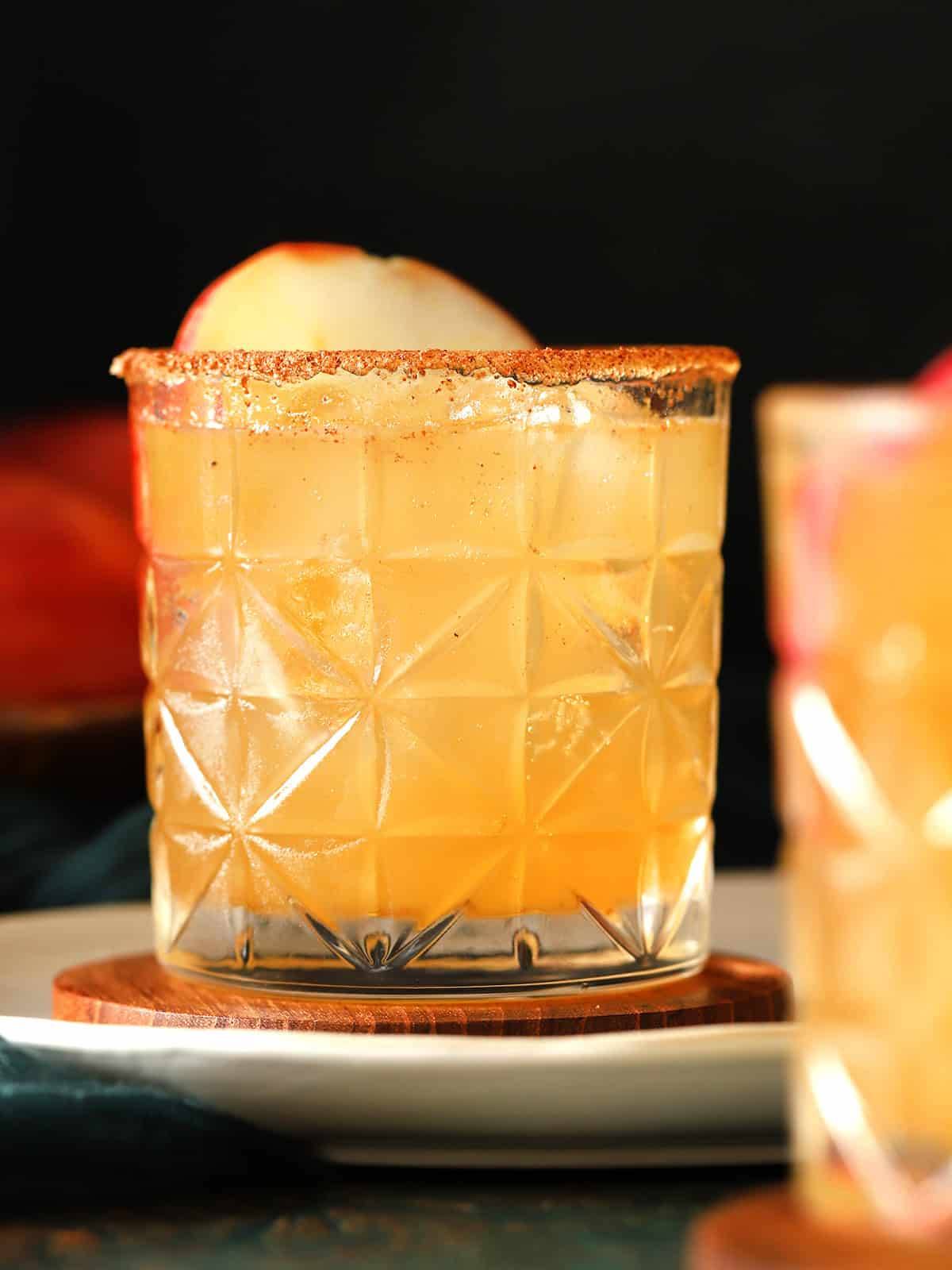 A pale yellow drink sits in a short glass garnished with an apple.