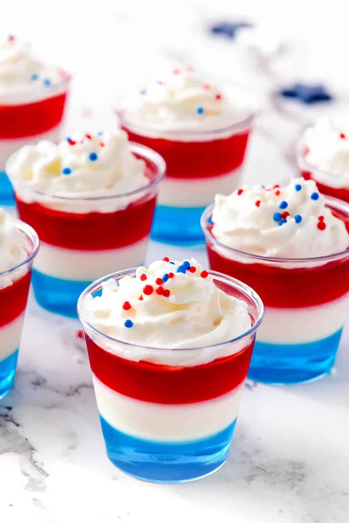 Layered red, white and blue jello shots topped with whipped cream and sprinkles sit on a white surface