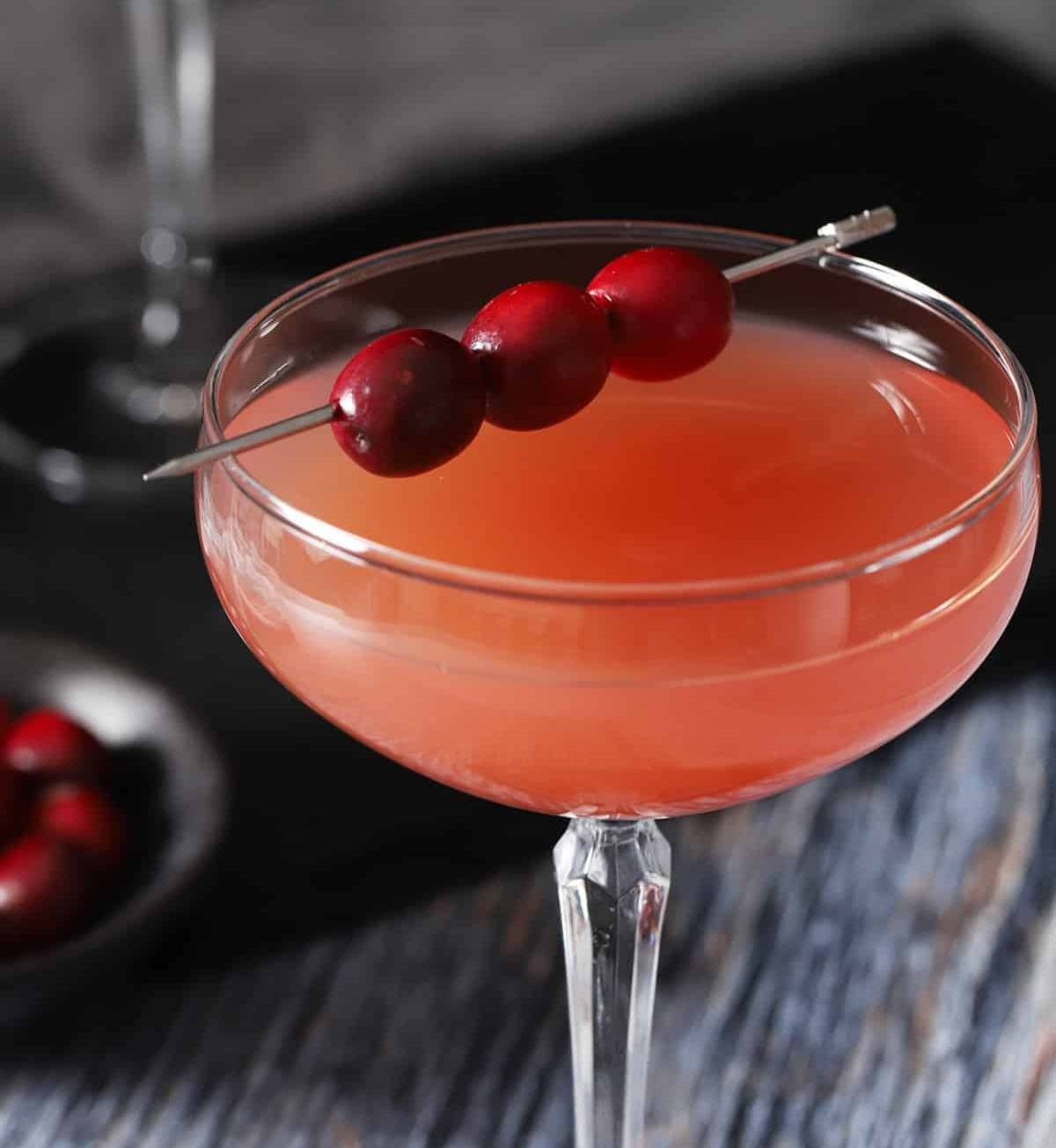A pink drink in a stemmed glass with a silver cocktail pick holding cranberries.