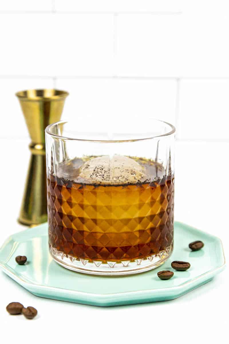 A rocks glass with a brown cocktail and a large sphere ice cube on a teal plate.