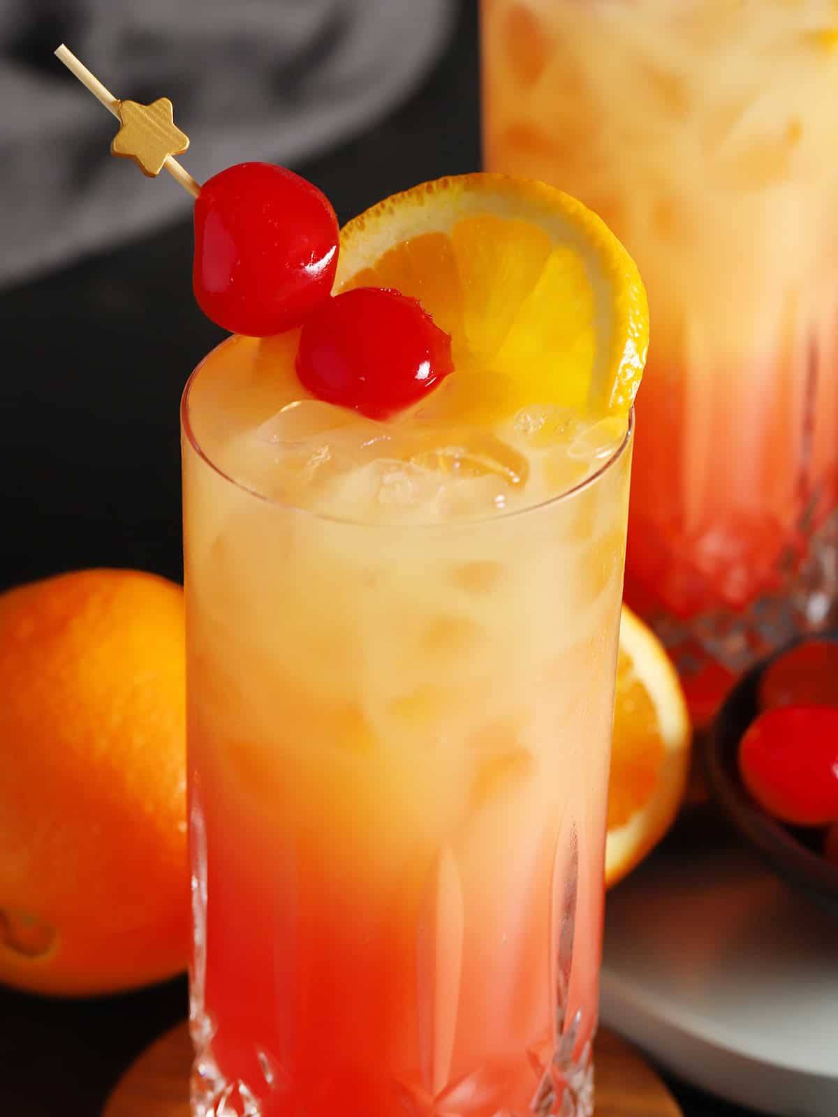 A cocktail that flows from red to orange garnished with cherries and an orange slice.