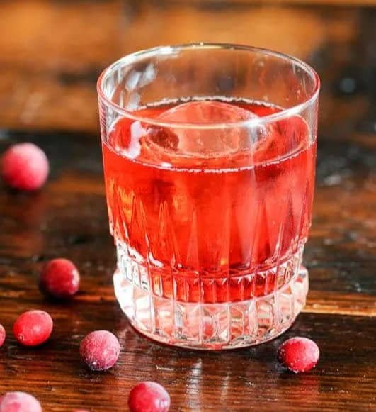 A red cocktail in a rocks glass with a large sphere ice cube near some cranberries.