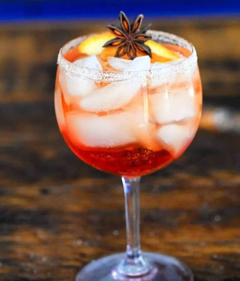 A red drink with plenty of ice in a stemmed glass topped with a star anise and orange slice.