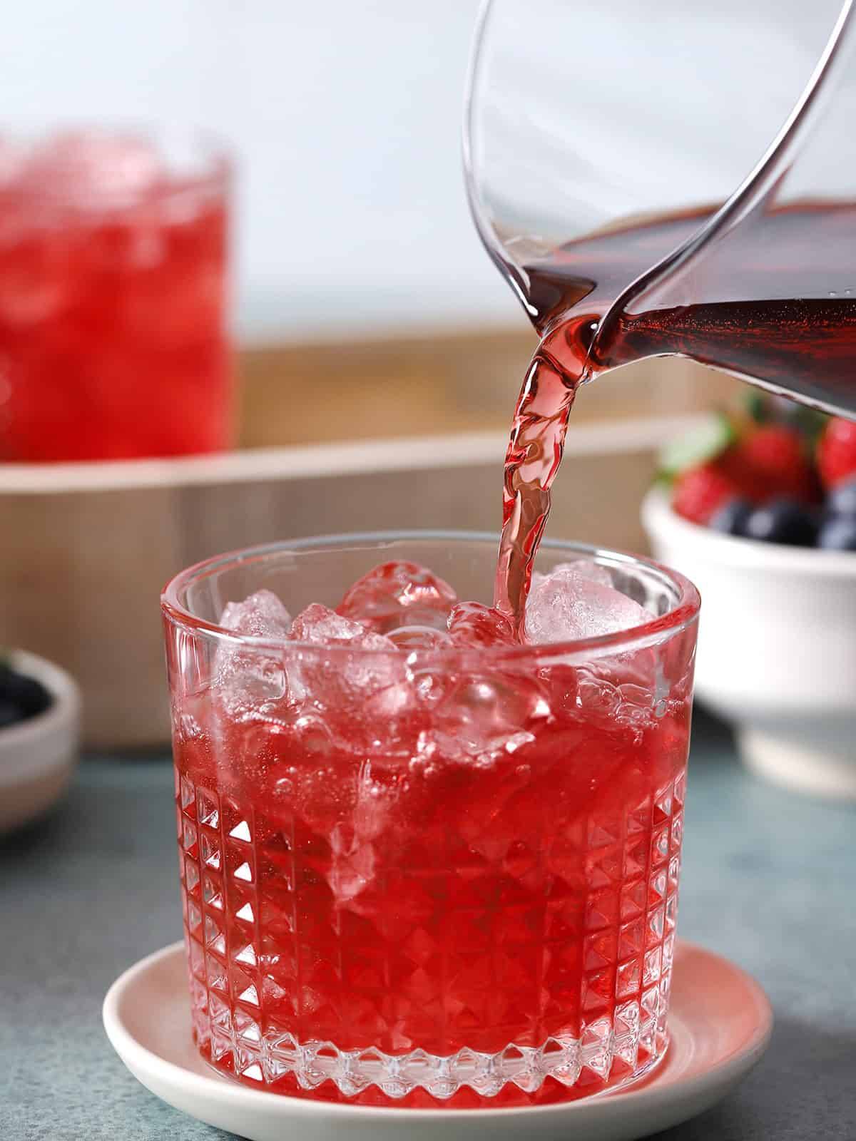 A red drink is poured from a clear glass pitcher into a rocks glass filled with ice.