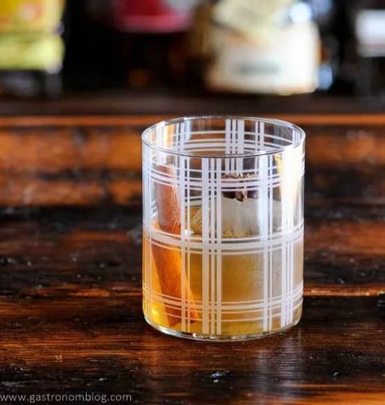 A light brown drink in a glass with a white plaid pattern sits on a wooden surface.