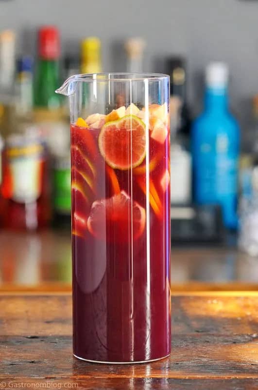 A tall glass pitcher without a handle holds a crimson liquid topped with fruit slices.