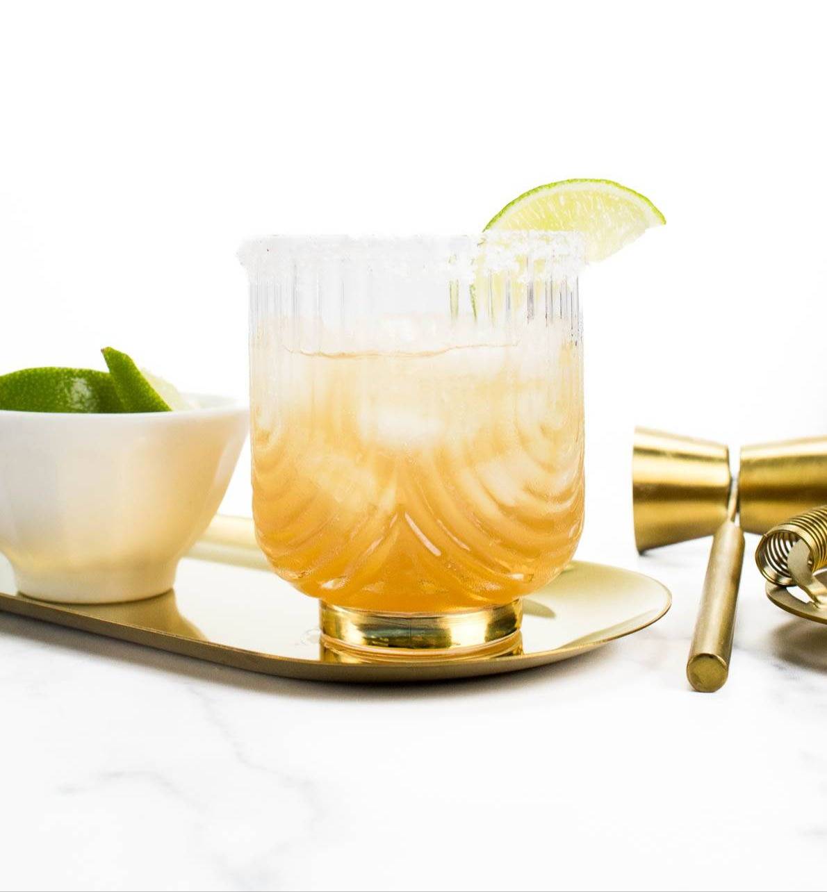 An amber drink in a glass rimmed with salt and a lime wedge sits on an oval gold tray near gold bar tools and a white bowl of lime slices.