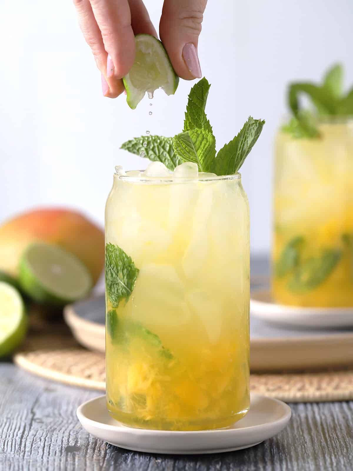 A hand squeezes a lime into a yellow drink filled with ice and mint leaves.