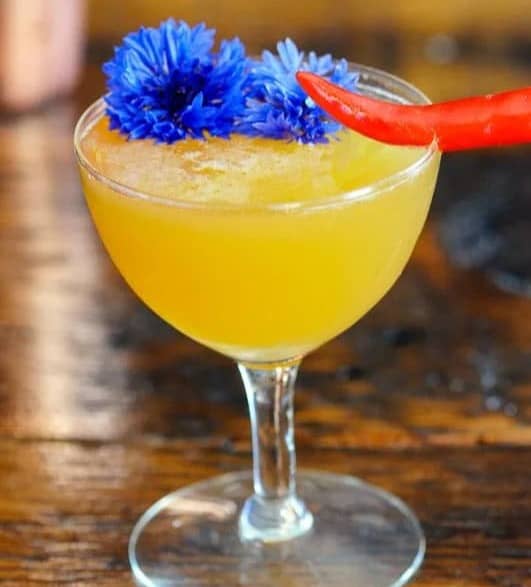 A yellow cocktail in a stemmed glass topped with blue flowers and a red pepper.