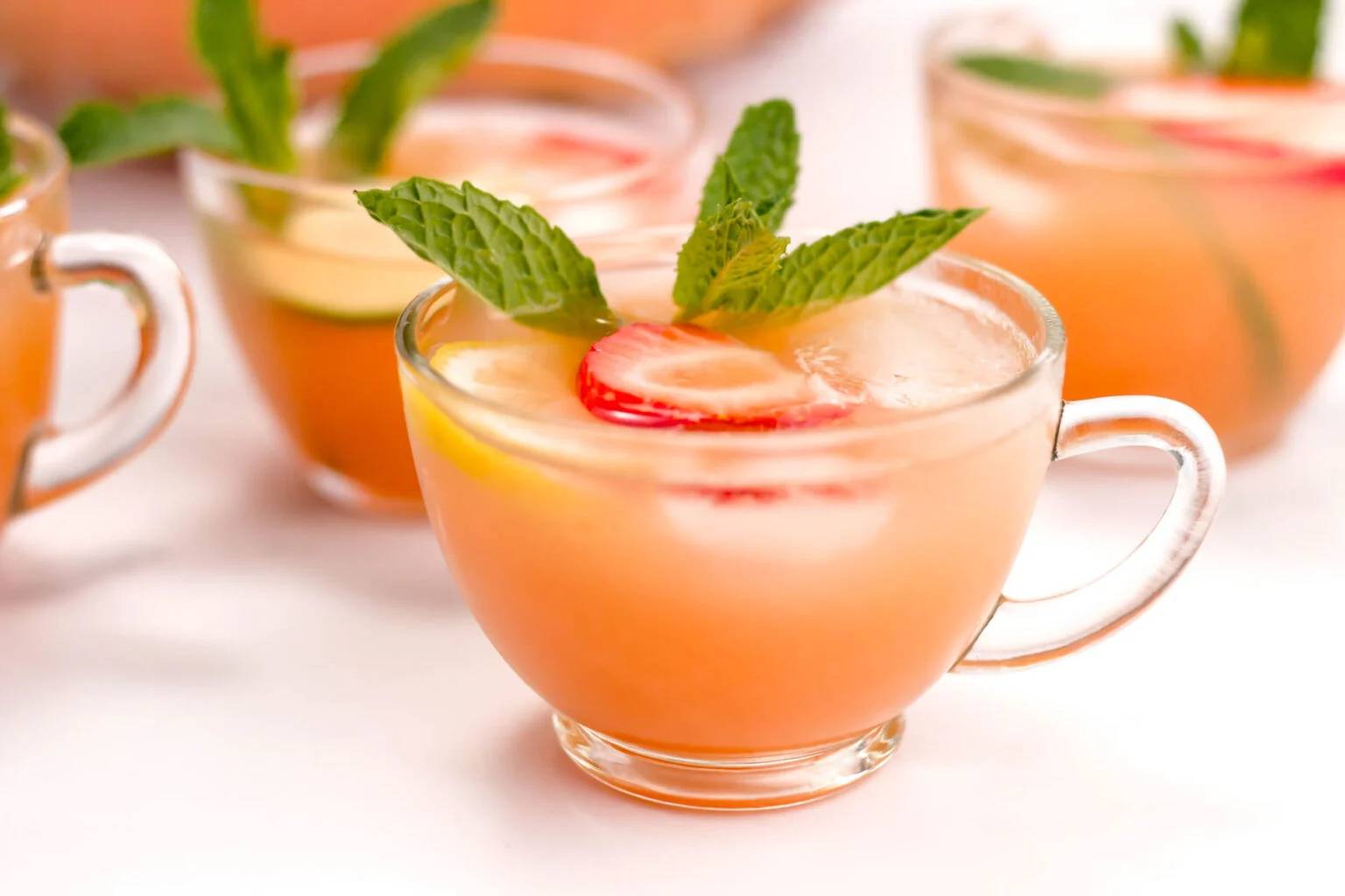 Clear glass teacups holding a pale pink drink garnished with fruit slices and a mint sprig.