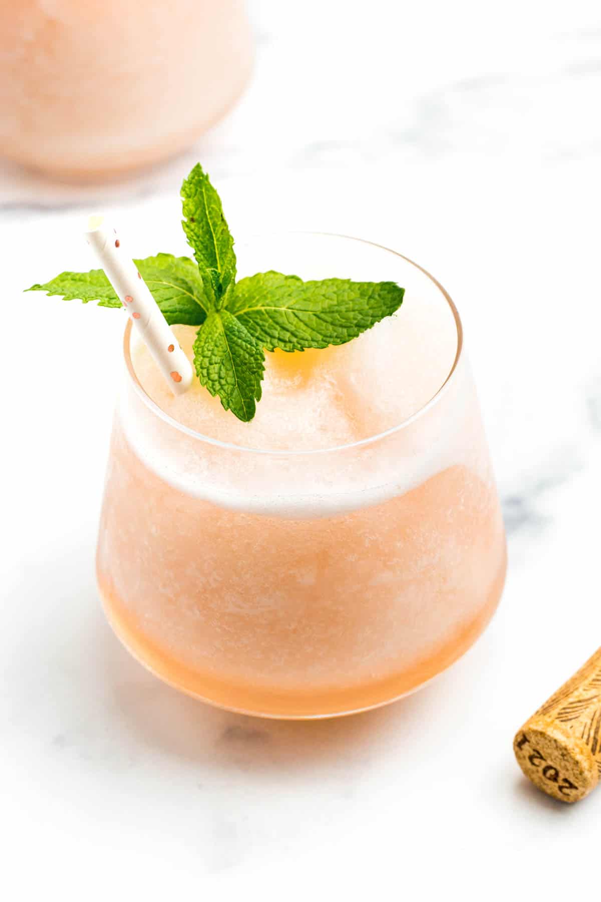 A stemless glass holding a light pink frozen cocktail topped with a sprig of mint and a white straw with rose gold dots.